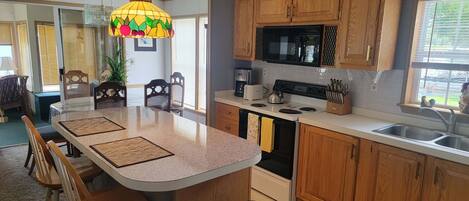 A kitchen with wooden cabinets, a central island with stools, a microwave, a stove, and a hanging fruit-themed light fixture.