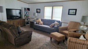 A living room with a grey sofa, striped armchairs, a brown armchair, a TV, a bookshelf, and two windows with blinds. - Cozy 2 bedroom Lake Cottage on Lamoka Lake in Bradford New York (Bradford)