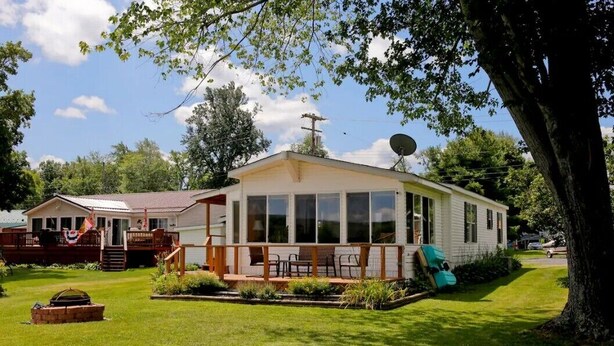 A single-story mobile home with a large front porch, a satellite dish, and a well-maintained lawn. - Cozy 2 bedroom Lake Cottage on Lamoka Lake in Bradford New York (Bradford)