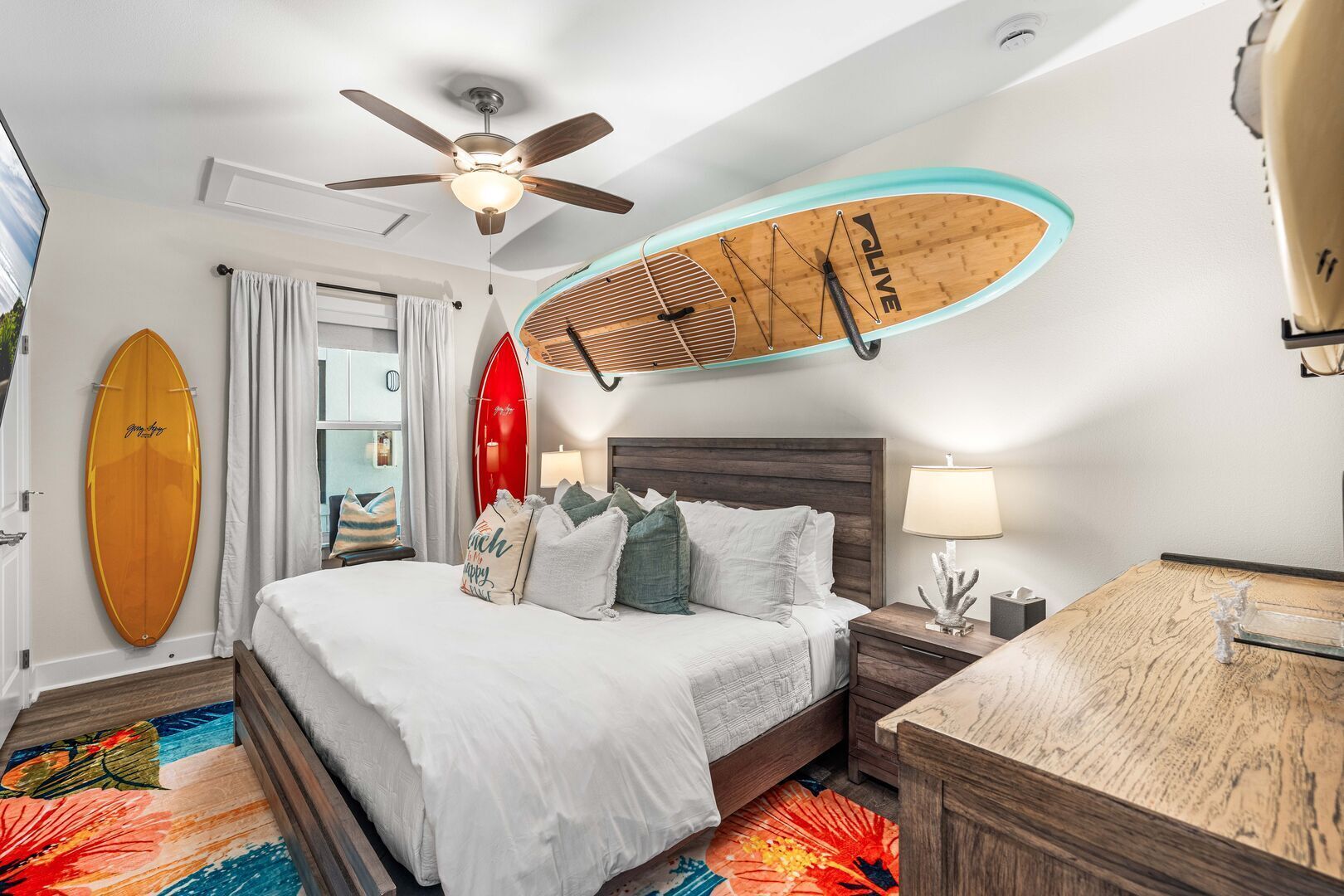 A hotel room with a bed, a ceiling fan, a surfboard, a nightstand, a desk, and a colorful rug.