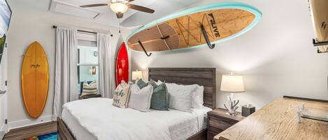 A hotel room with a bed, a ceiling fan, a surfboard, a nightstand, a desk, and a colorful rug.