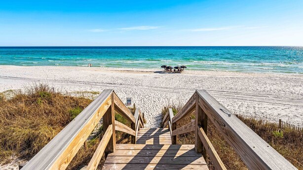 Un paseo marítimo de madera que conduce a una playa con agua azul cristalina y algunas sombrillas a lo lejos.