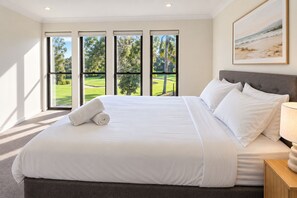 A large bed with white linens, a grey headboard, and a view of a green landscape through the windows.