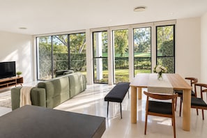 A modern living room with a large window, a flat-screen TV, a green sofa, a dining table with chairs, and a bench.