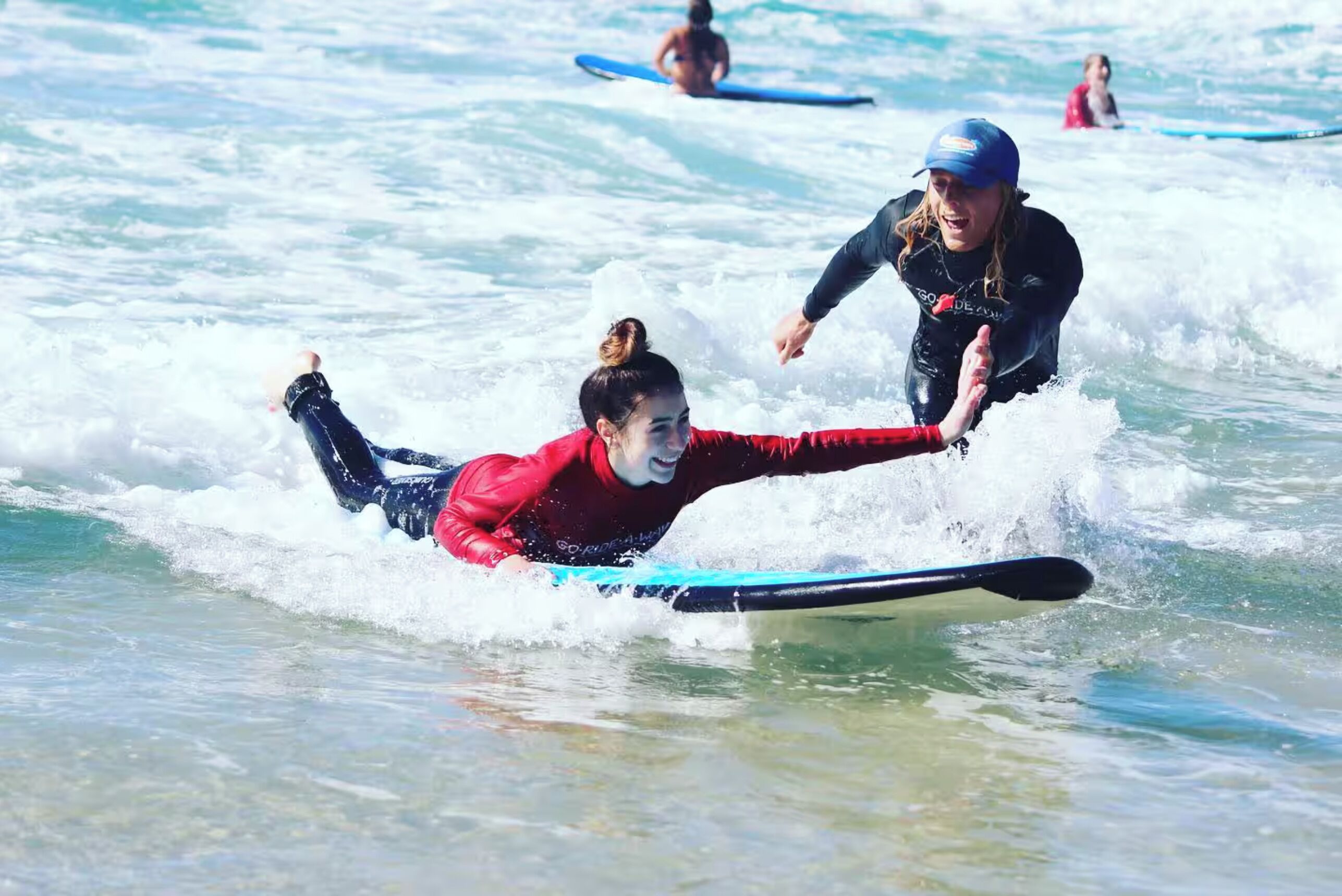 A person in a red wetsuit on a surfboard in the ocean, with another person in a blue wetsuit nearby.