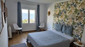 Ein Schlafzimmer mit einem Bett, zwei Stühlen, einem Fenster mit Vorhängen, tropischen Tapeten und Blick aufs Meer.