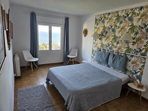 A bedroom with a bed, two chairs, a window with curtains, a tropical wallpaper, and a view of the sea. - LOS TORRINOS Dream Villa with Mediterranean Charm (Roses)