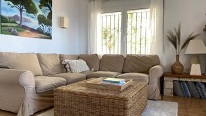 Ein Wohnzimmer mit einem beigen Sofa, einem Korbtisch und einem Baumgemälde an der Wand.