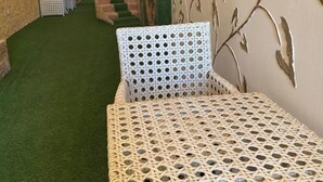 Una mesa y sillas de mimbre en un espacio exterior cubierto con césped artificial, paredes de ladrillo y una pared decorativa con motivos de hojas.