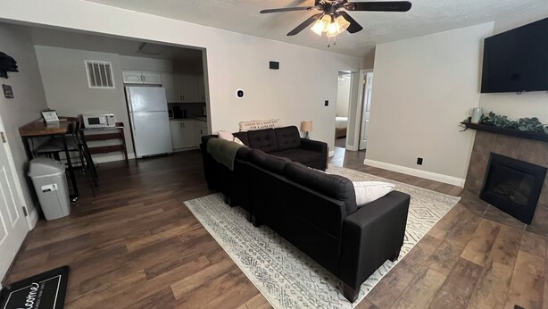 A living room with a dark sofa, a patterned rug, a ceiling fan, a fireplace, and a kitchen area in the background. - Convenient & Cozy condo on 400 (Logan)