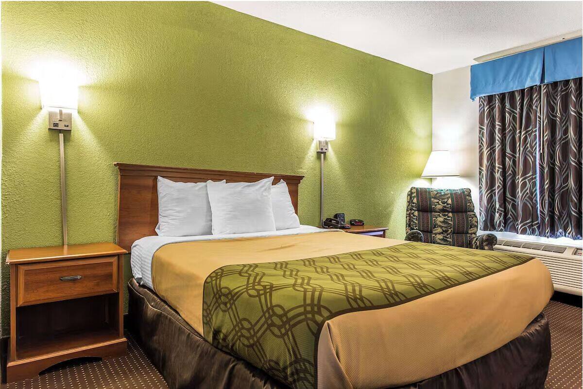 Una habitación de hotel con pared verde, cabecera de madera, cama con almohadas blancas, cubrecama estampado, mesita de noche, lámpara, cortina estampada y ventana con persianas.