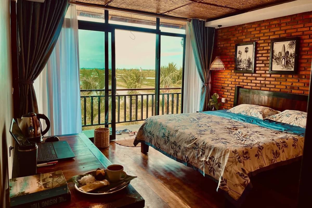 A bedroom with a large bed, a laptop on a wooden table, and a view of greenery outside.