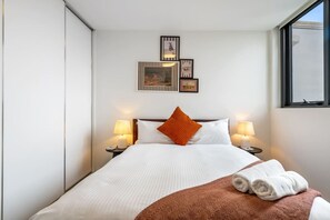 A hotel room with a bed, two bedside lamps, a window, and framed artwork on the wall. - Urban Cowboy Loft | Skyline Views, Pool & Parking (St Kilda)