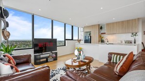 Una sala de estar moderna con un televisor grande, sofás de cuero, una mesa de centro y vistas de la ciudad a través de amplias ventanas.