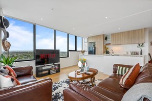 A modern living room with a large TV, leather sofas, a coffee table, and a view of the city through large windows. - Urban Cowboy Loft | Skyline Views, Pool & Parking (St Kilda)