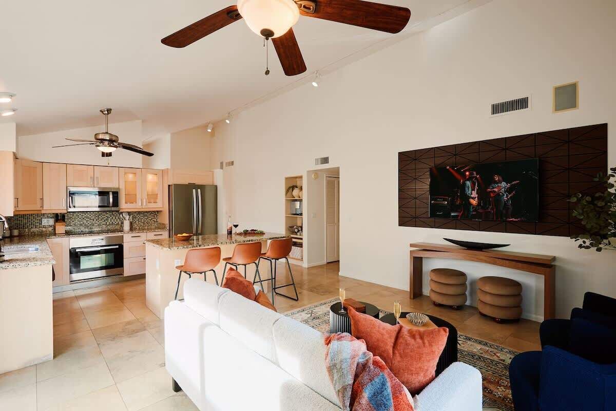 A modern kitchen with a dining area, a living room with a sofa, and a TV mounted on the wall.