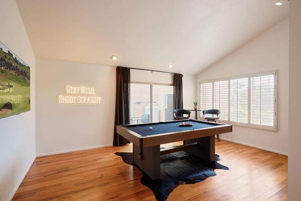 A room with a pool table, a large window with blinds, and a wall with a motivational quote.