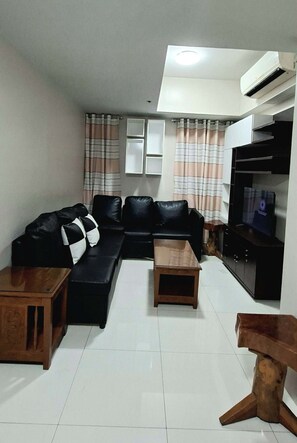 A living room with a black sofa, two wooden side tables, a TV on a cabinet, and striped curtains. - Cozy Modern 2-bedroom condo with AC in lovely Newport City (Pasay City)