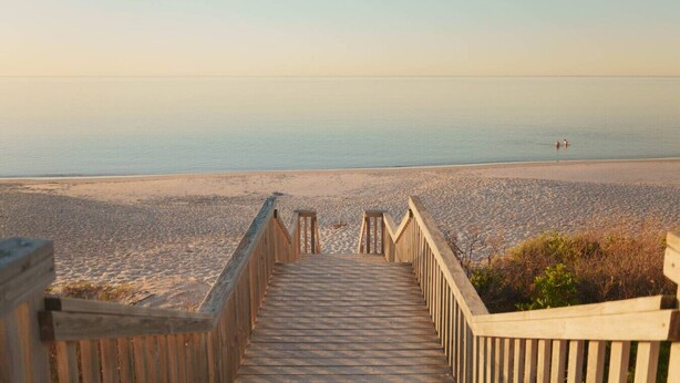 Une promenade en bois qui mène à une plage o ù la mer est calme et le ciel dégagé.