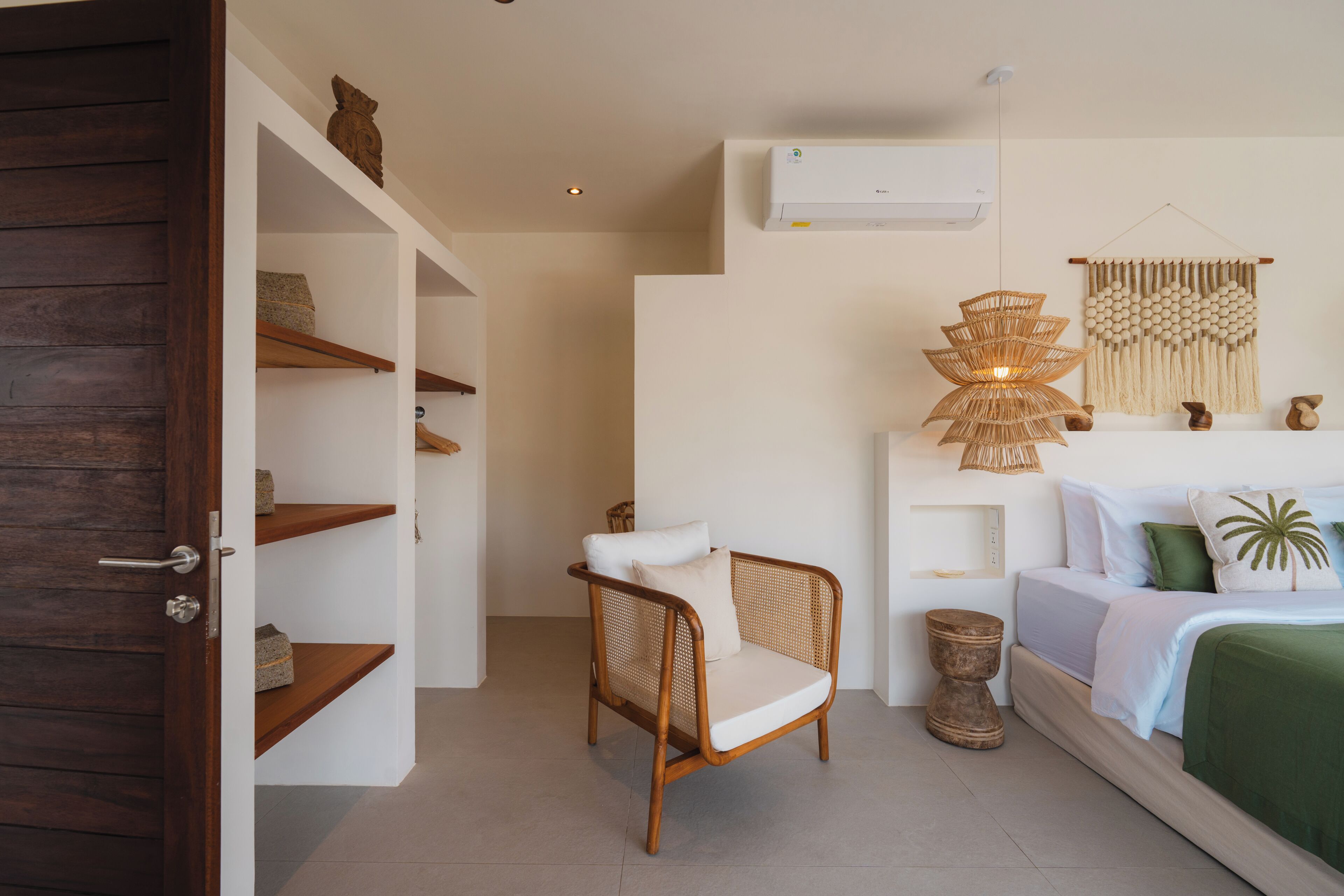 A bedroom with a bed, a wicker chair, a wall-mounted air conditioner, and a decorative wall hanging.