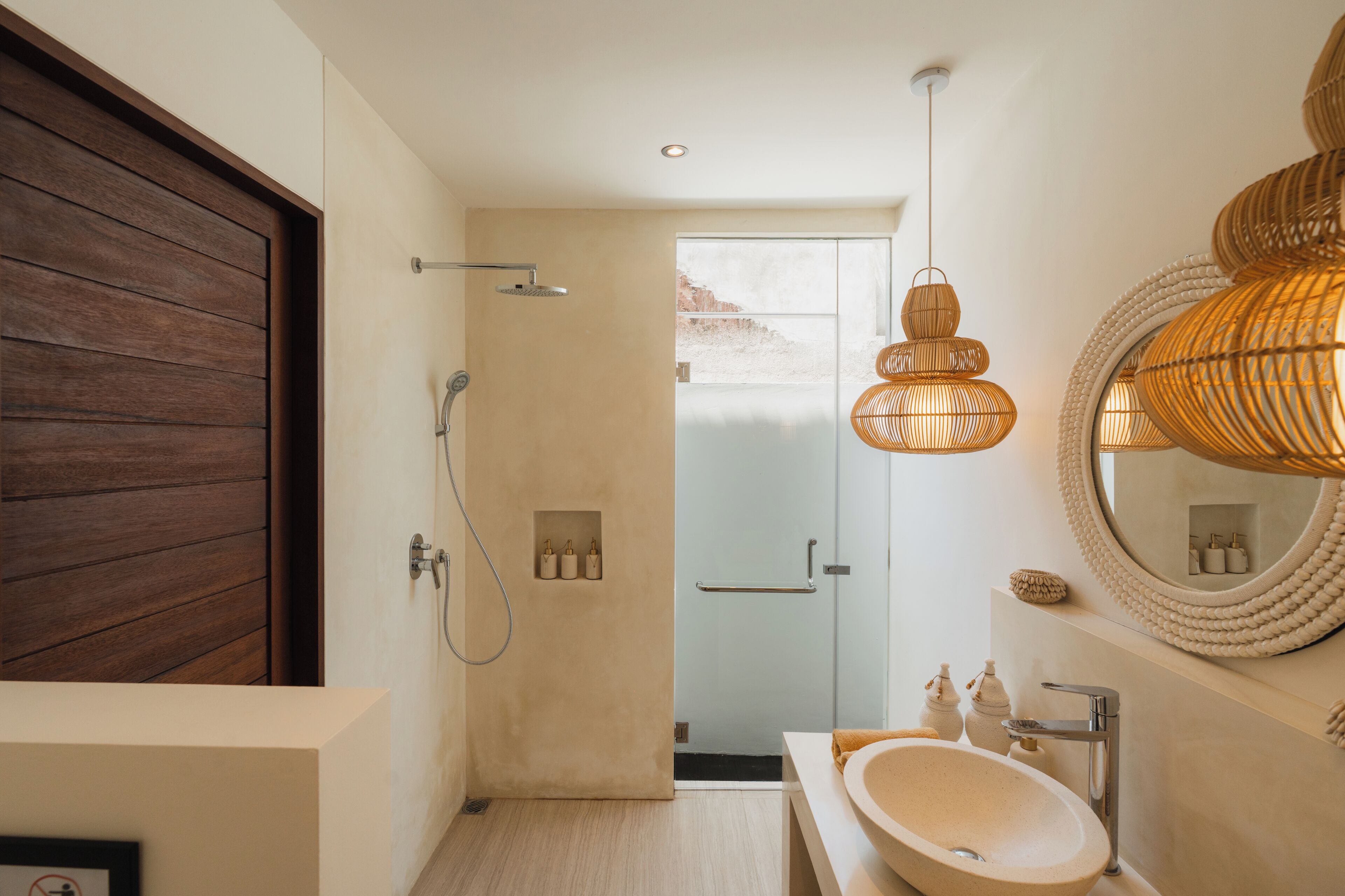 A modern bathroom with a wooden door, a glass shower enclosure, a round mirror, and a white sink.