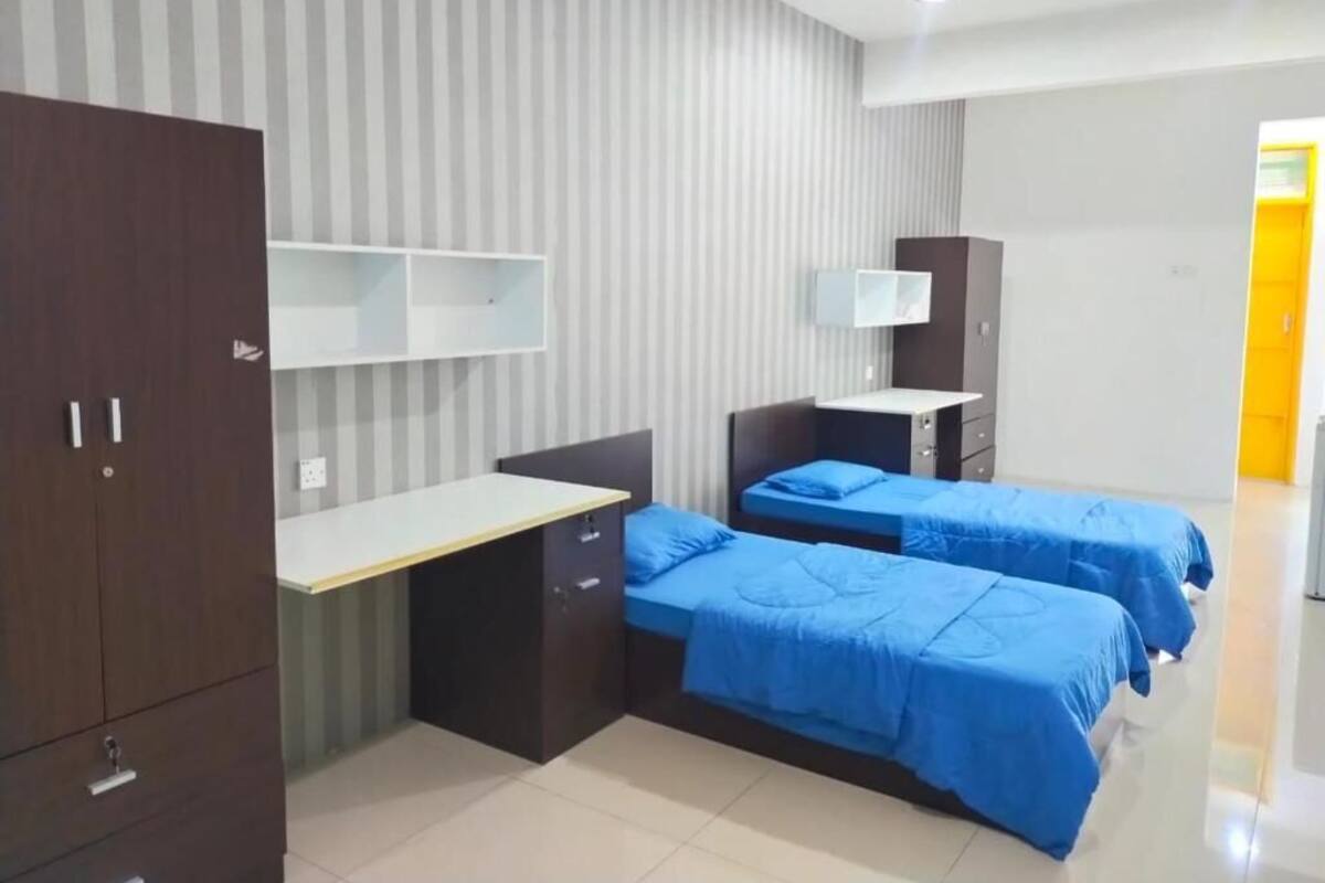 A room with a desk, two beds with blue bedding, a wardrobe, and a shelf unit.