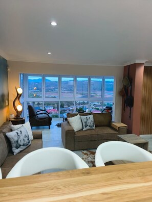 A spacious living room with a large window offering a view of a hilly landscape. There is a brown sofa with decorative pillows, a black chair, and a white armchair. A wooden coffee table is in the foreground.