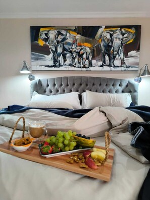 A neatly made bed with a grey headboard, a tray with breakfast items including grapes, nuts, and a drink, and a painting of abstract figures above the bed.