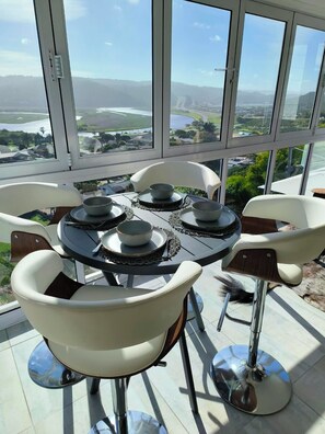 Un coin repas avec une table dressée pour quatre personnes, entouré de chaises blanches aux pieds chromés, donnant sur un paysage pittoresque.