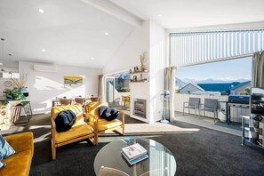 A modern living room with a sofa, a coffee table, and a dining area with chairs. There is a fireplace and a view of the mountains through the window. - Generous Townhouse: Views & Pool (Wanaka)