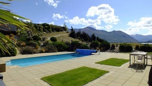 A swimming pool with a view of mountains, a patio with a table and chairs, and a well-maintained lawn.