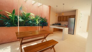 A modern kitchen with a wooden dining table and bench, a built-in plant wall, and a kitchen island with a refrigerator. - La Garza, View of the interior garden. (Mérida)