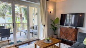 A living room with a sofa, a wooden coffee table, a TV, and a sliding glass door leading to an outdoor area with a patio and plants. - Poipu 3BR/3BA corner villa, poolside, A/C (Koloa)