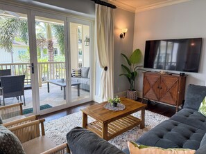 Ein Wohnzimmer mit Sofa, Couchtisch aus Holz, Fernseher und einer Glastür zum Gartenbereich mit Terrasse und Pflanzen.
