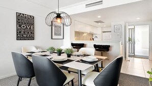 A modern dining area with a white table and black chairs, a pendant light, and a kitchen in the background. - Balcony Views, Parking, Pool & Gym | Walk to CBD (Spring Hill)