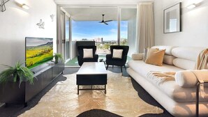 A modern living room with a sofa, a coffee table, and a TV on a stand. There is a balcony with a view of the city and a ceiling fan. - Balcony Views, Parking, Pool & Gym | Walk to CBD (Spring Hill)