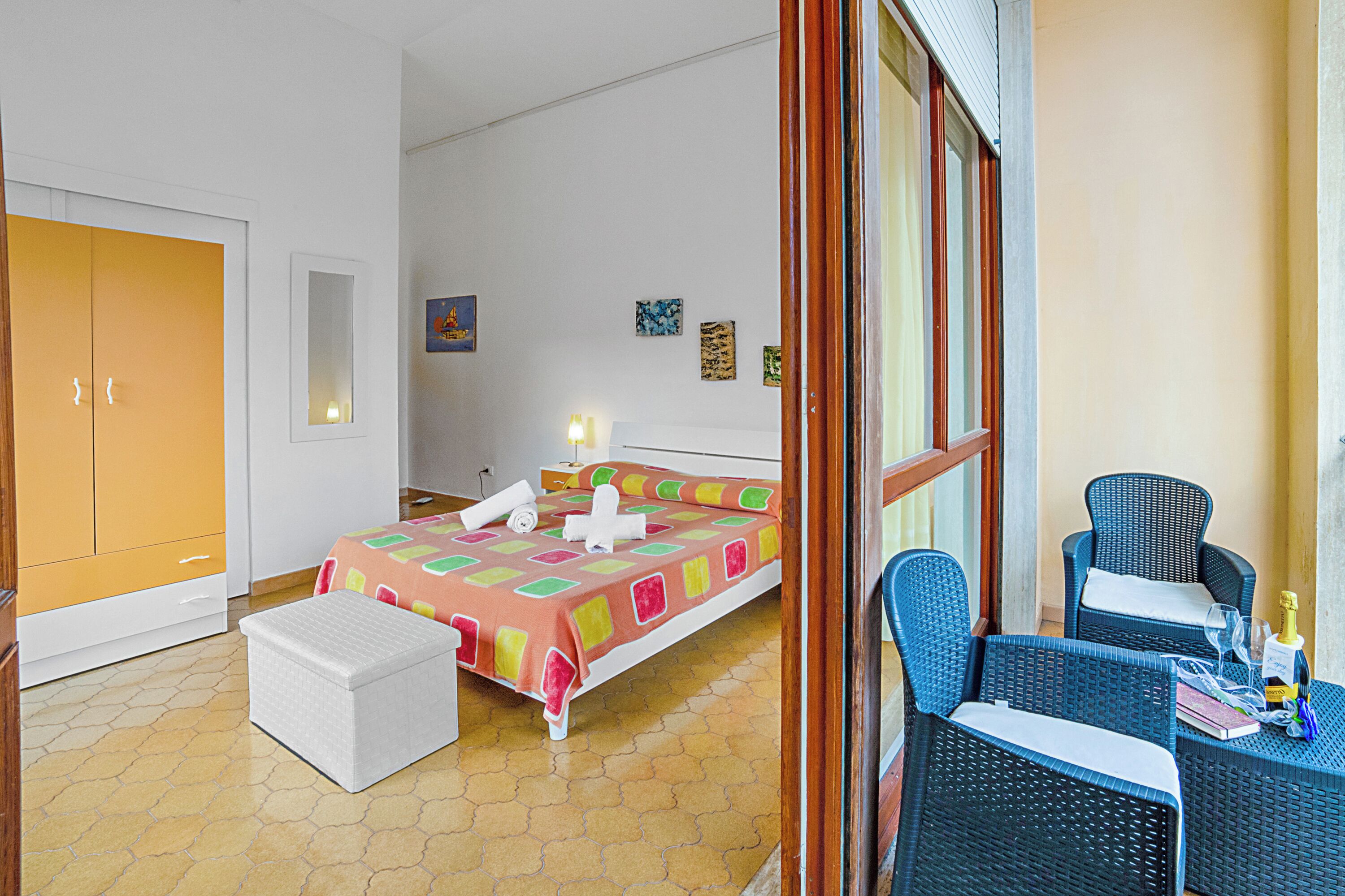 A bedroom with a bed, a white chest of drawers, a wicker chair, and a small table with a bottle and glasses.