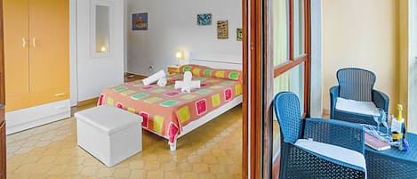 Un dormitorio con una cama, un aparador blanco, una silla de mimbre y una mesita con una botella y vasos.