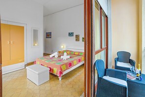 Ein Schlafzimmer mit einem Bett, einer weißen Kommode, einem Korbstuhl und einem kleinen Tisch mit einer Flasche und Gläsern.