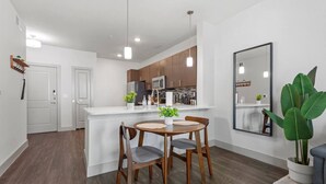 A modern kitchen with wooden cabinets, a white countertop, and pendant lighting. There is a dining area with a wooden table and chairs, and a large potted plant.