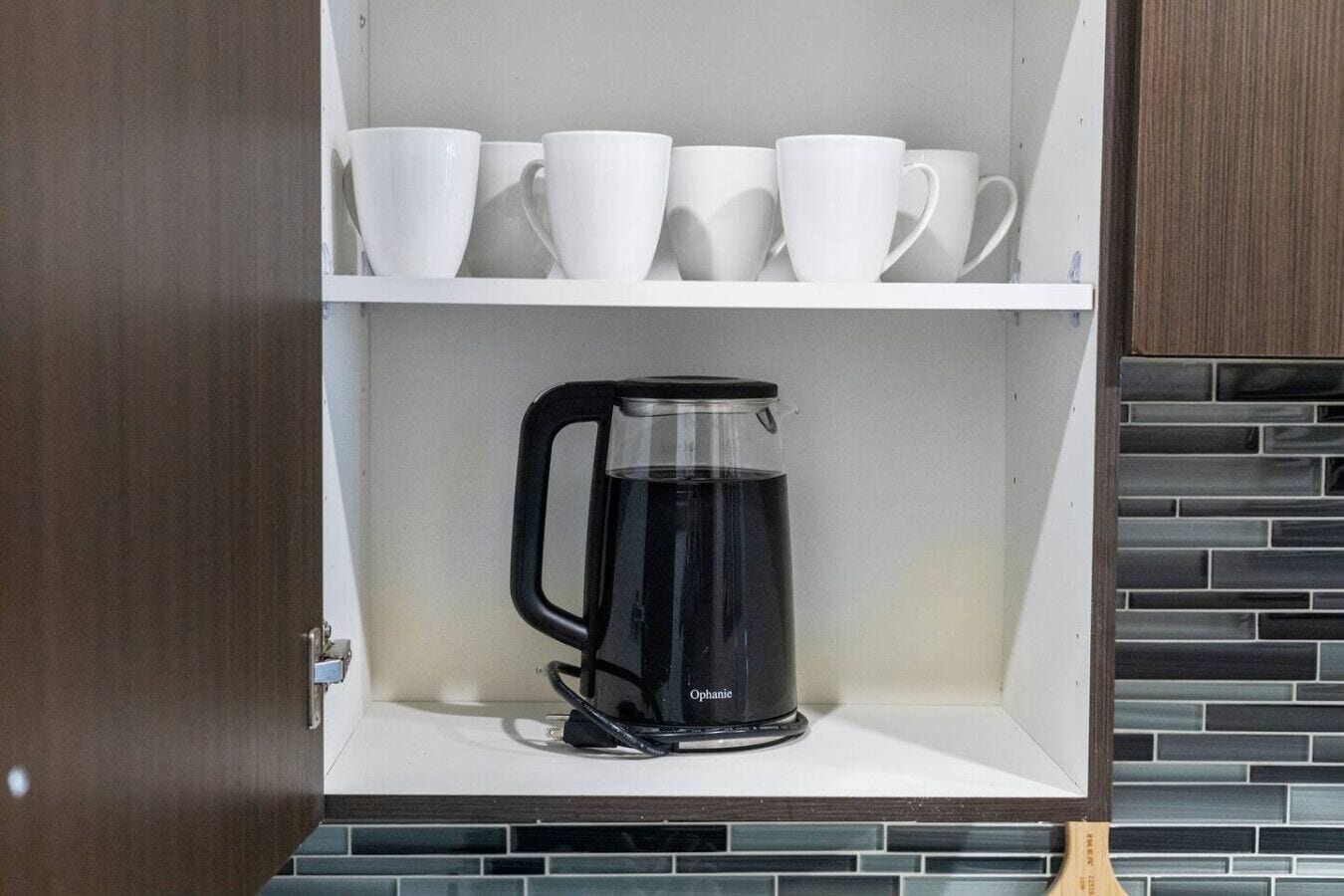 A kitchen with a coffee maker on the counter, white cups on the shelf above, and a tiled backsplash.