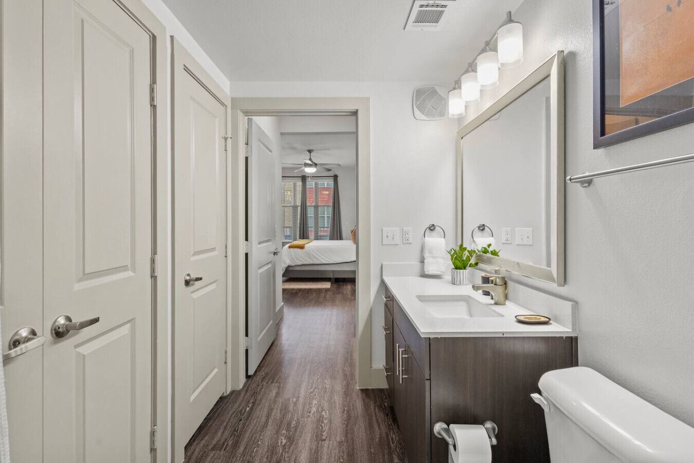A narrow hallway leading to a bedroom with a bed, a bathroom with a sink, mirror, and towel rack.