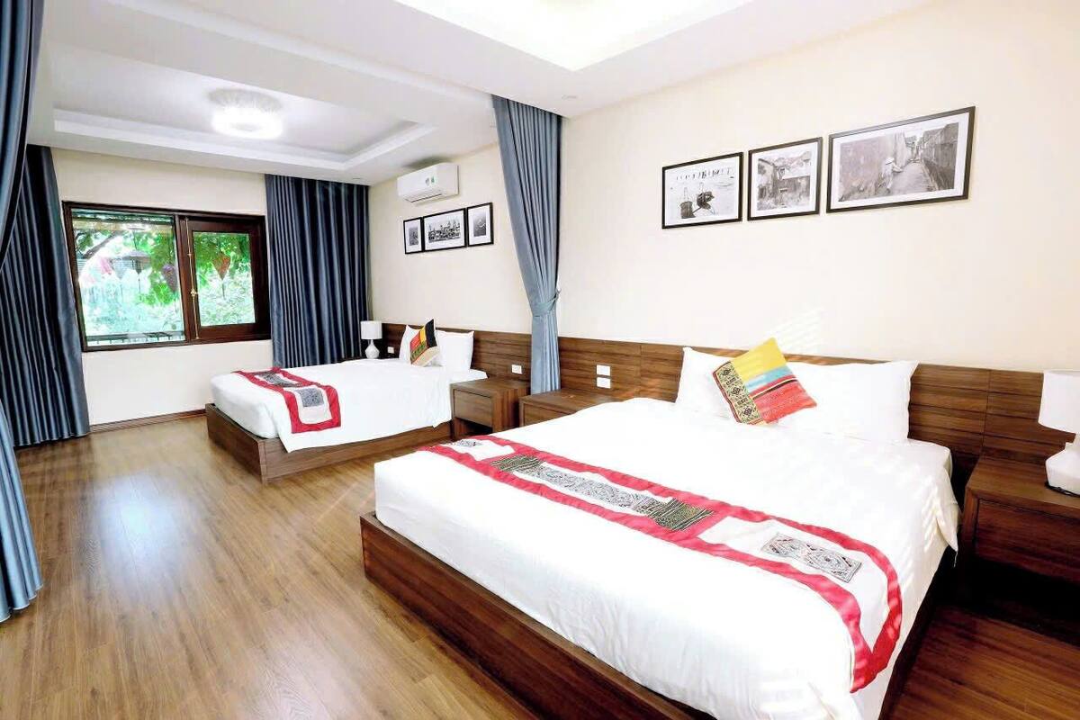 Habitación de hotel con una cama grande, una cama más pequeña, piso de madera, una ventana con cortinas e imágenes enmarcadas en la pared.
