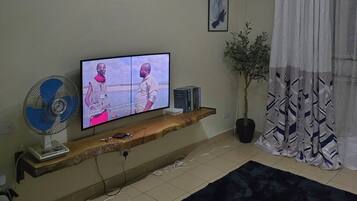 Una sala de estar con televisión, ventilador, planta y ventana con cortinas.