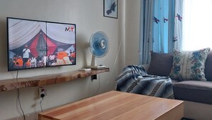 Una sala de estar con un televisor montado en la pared, un ventilador, un sofá y una mesa de madera con ruedas.