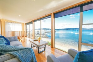 A spacious living area with a large window overlooking a beach and sea, a wooden floor, and a balcony with a railing. - StayAU Riverfront Paradise Pet friendly (Beauty Point)