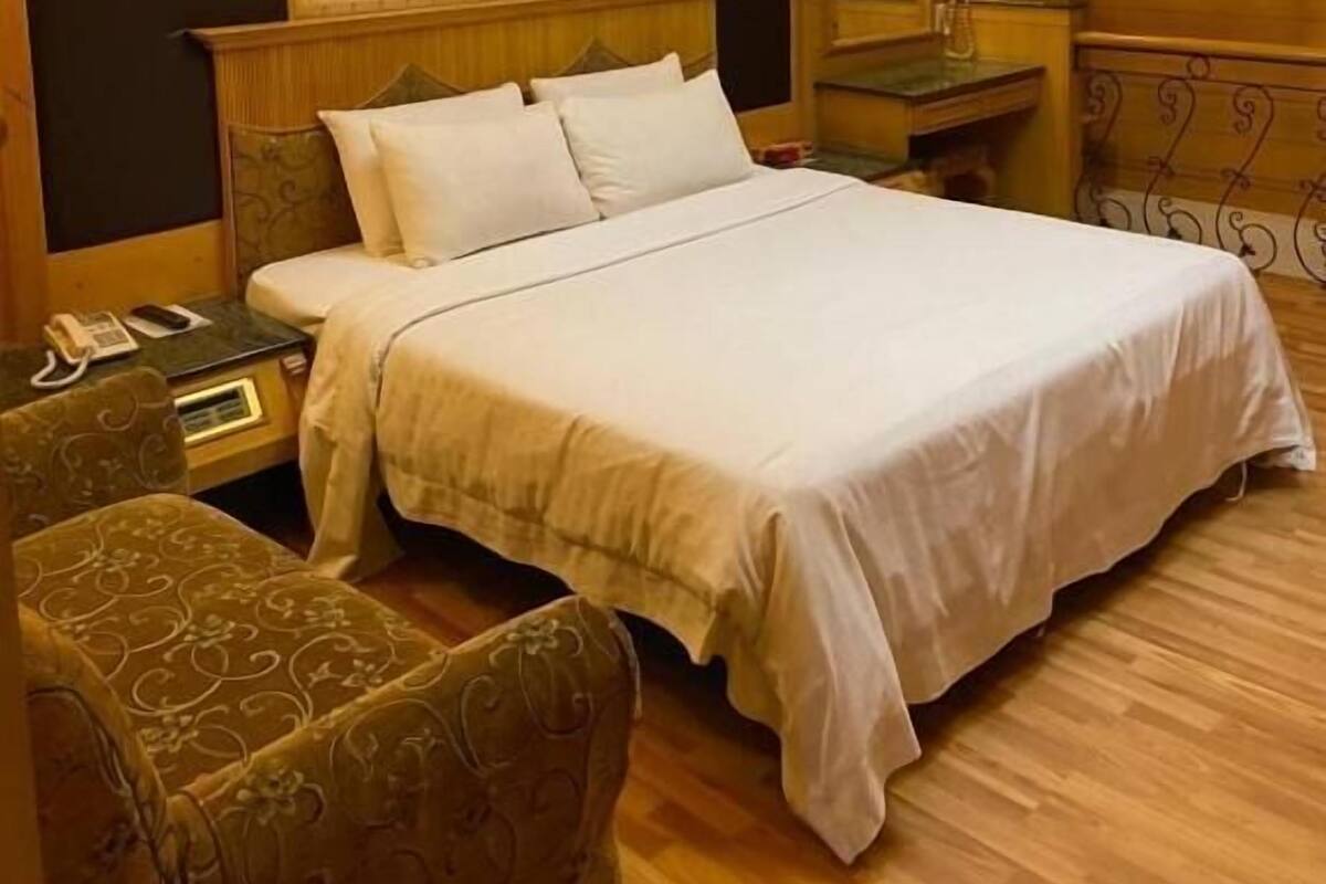 A hotel room with a large bed, a bedside table with a lamp, a chair, and a wooden ceiling with a skylight.