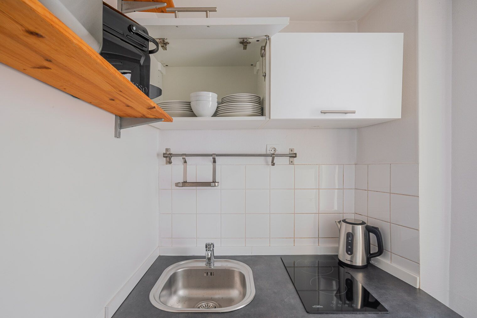 A compact kitchen with a built-in sink, a kettle, and a shelf above the sink.