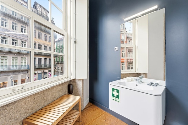 Une salle de bain moderne avec une grande fenêtre, un lavabo blanc orné d’une croix verte et un banc en bois.