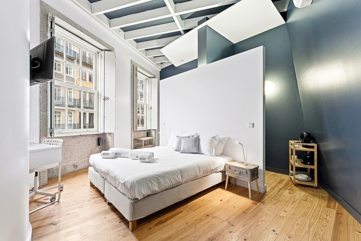 Ein modernes Schlafzimmer mit einem großen Bett, einem Schreibtisch und einem Stuhl, einem Fenster mit Blick auf Gebäude und einer dunkel gestrichenen Akzentwand.
